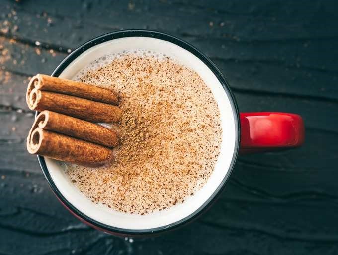 A frothy beverage in a red mug topped with a sprinkle of cinnamon powder and adorned with cinnamon sticks on a dark wooden background.