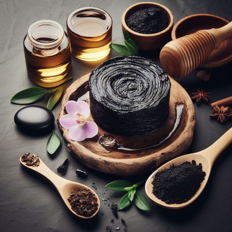A black swirl of Shilajit resin on a wooden plate surrounded by small jars of golden liquid, wooden spoons filled with powdered and granulated forms, and aromatic embellishments like a pink orchid, star anise, and fresh green leaves on a slate background.
