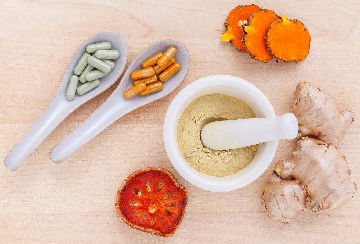 Green and orange capsules in ceramic spoons, sliced turmeric root, dried bael fruit slice, and ground herbal powder in a mortar with pestle on a wooden background.