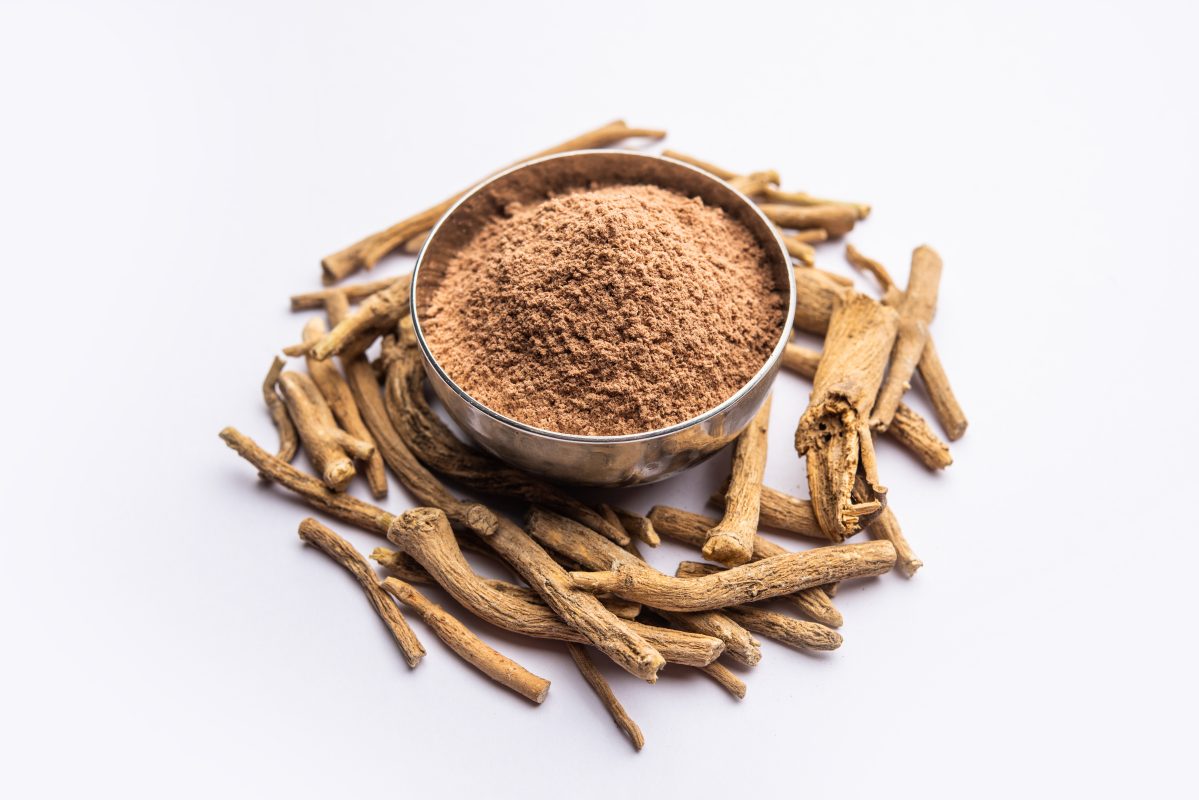 Bowl of finely ground Ashwagandha powder surrounded by dried Ashwagandha roots on a white background.